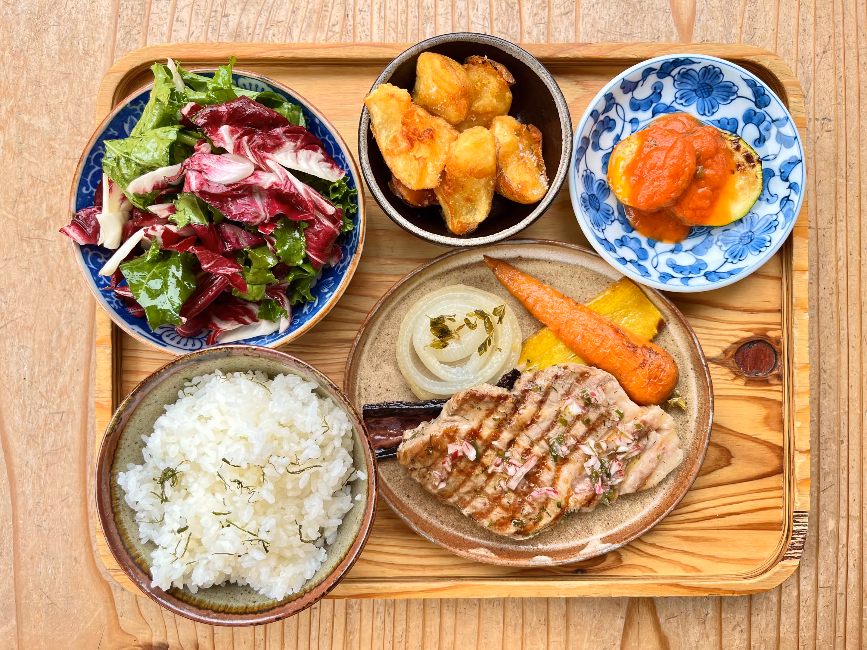 A colorful lunch plate served on a wooden tray. Grilled meats, potatoes, salad, rice, and other items are arranged on the tray.