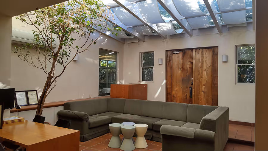 The waiting room has a relaxed atmosphere with an open ceiling and wooden doors.Gray sofas are arranged and small wooden tables are placed.