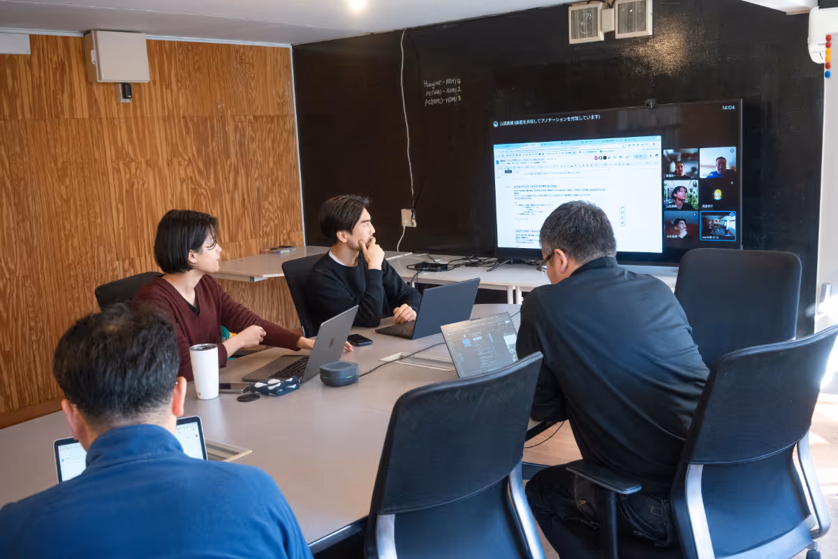 Four businesspeople with serious expressions face the computer. On the wall is a large monitor showing the online meeting.
