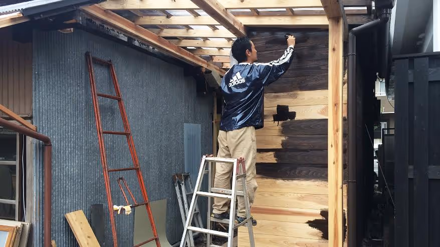 Employees apply dark brown paint to the wooden exterior of an old building.