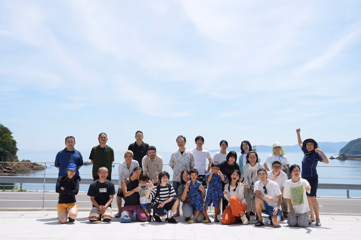 Group photo of the 2024 Suo Oshima company trip. Everyone is lined up with smiles on their faces, with the big sea and sky in the background.