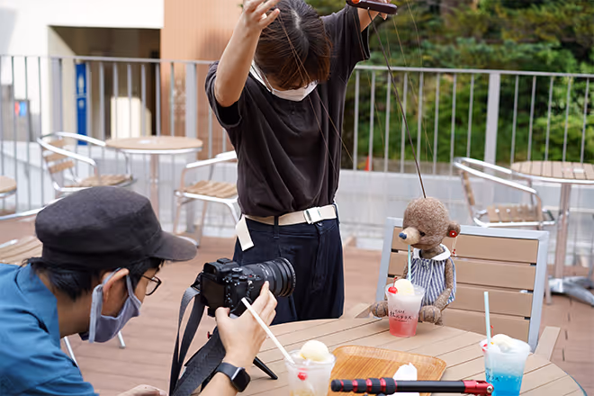 A person is outdoors, manipulating a stuffed animal for a photo shoot. The stuffed animal is drinking a drink.