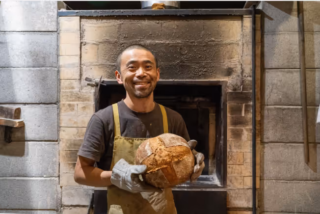 Here is a photo of Shiomi, the smiling baker, holding a loaf of bread baked in front of his stone oven with both hands.
