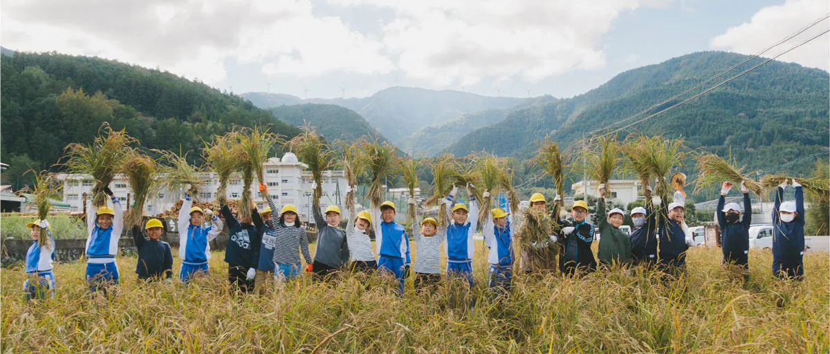 This is a picture of children experiencing rice harvesting, standing smiling as they hold their harvested rice high in the air. The mountains are in the background.