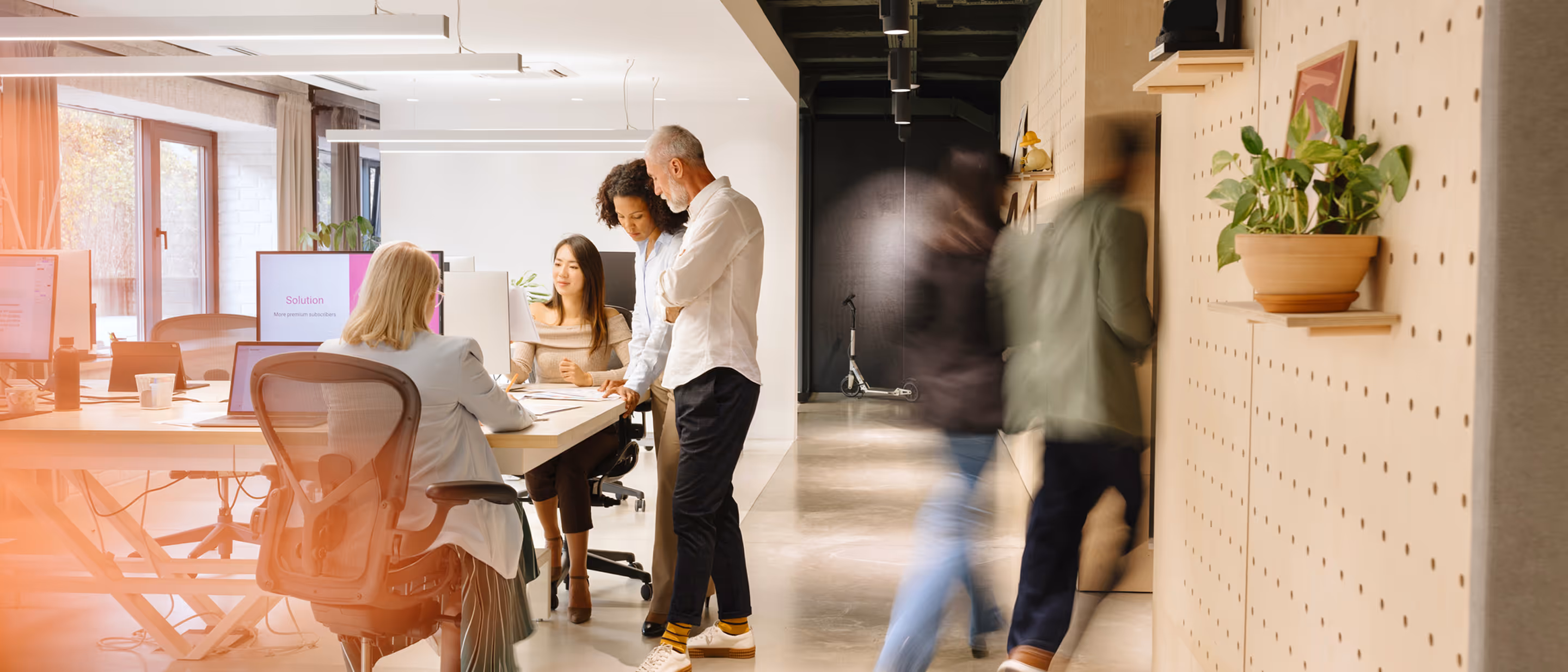 Bright office scene. In the foreground, a woman works on a computer at her desk. In the background, three business professionals are standing and talking. Further back in the hallway, two figures are seen in motion.